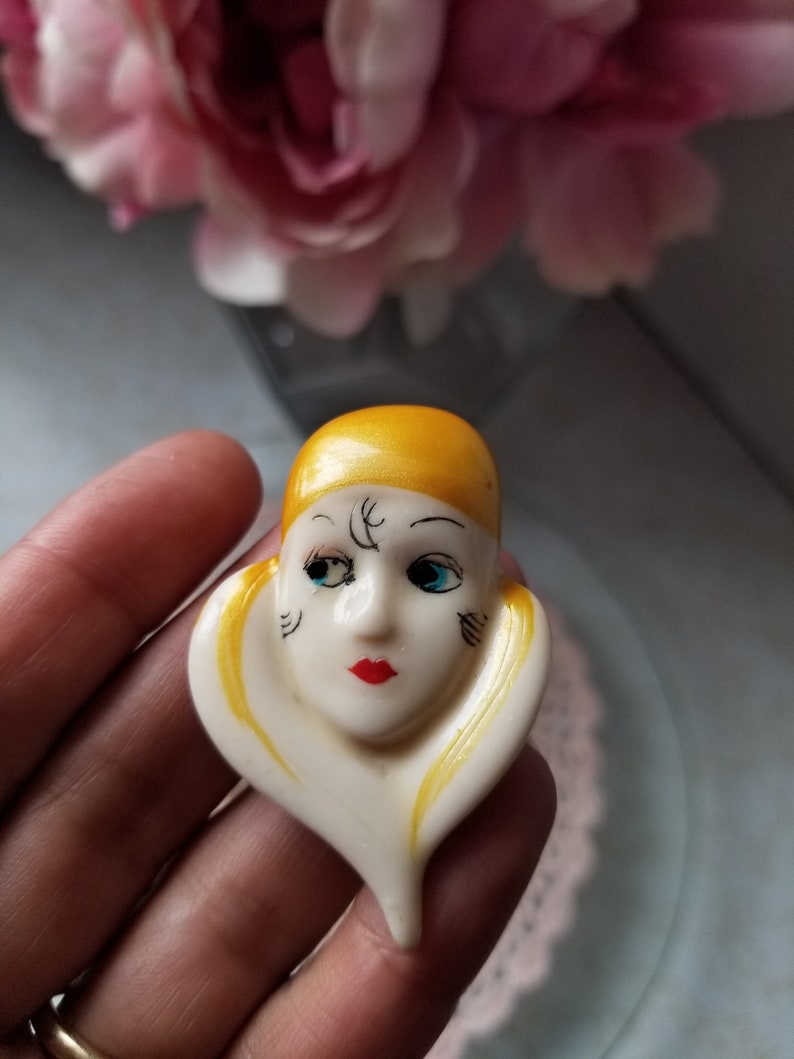 K&ouml;nnte beinhalten: Eine wei&szlig;e Keramikbrosche mit einem bemalten Gesicht und gelben Haaren. Das Gesicht hat schwarze Augen, einen roten Mund und schwarze Markierungen.