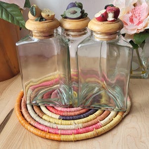 May include: Three glass jars with wooden lids and fruit-shaped tops. The jars are sitting on a colorful woven mat.