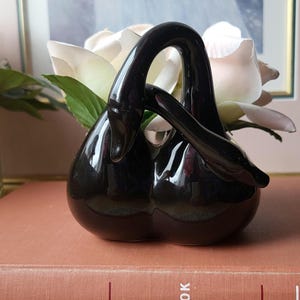 Könnte beinhalten: Eine glänzend schwarze, schwanförmige Keramikvase mit weißen Rosen und Grünzeug. Die Vase steht auf einem Vintage-Buch mit dem Titel "The Modern Handbook of Humor".