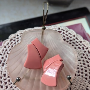 Pode incluir: Um prato vintage rosa em forma de concha com uma alça de metal e dois brincos rosa abstratos. Os brincos têm um design curvo. O prato está sobre uma toalha de crochê branca.