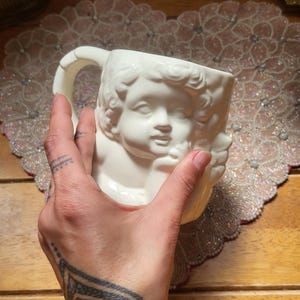 May include: White ceramic mug featuring a cherub face design. The mug has a handle and is held by a hand. The mug is on a decorative beaded placemat and wooden surface.