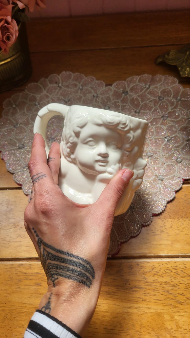 May include: White ceramic mug featuring a cherub face design. The mug has a handle and is held by a hand. The mug is on a decorative beaded placemat and wooden surface.