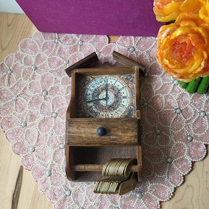 May include: A wooden wall clock with a drawer and a key holder. The clock face has Roman numerals and decorative details. The clock is sitting on a pink beaded placemat, with a purple book and orange flowers in the background.