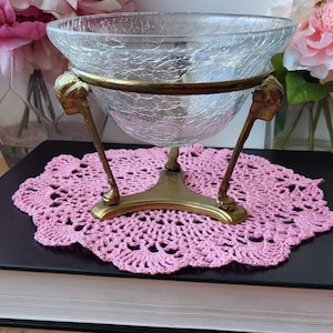 Puede incluir: Un cuenco de vidrio transparente con un acabado craquelado se asienta sobre un soporte de metal dorado con tres patas. El soporte tiene un diseño decorativo con patas en forma de garras.