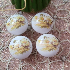 Peut inclure: Quatre boutons de meuble blancs avec un motif floral de marguerites et de blé. Les boutons sont disposés sur une surface perlée rose, avec un élément décoratif vert en arrière-plan.