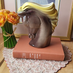 Op de afbeelding: Een keramieken paardenkopfiguur, bruin met een gele manen, rustend op een vintage boek getiteld "The Modern Handbook of Humor". De figuur staat op een roze kanten kleedje.