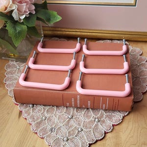 Puede incluir: Seis tiradores de armario rosas con tornillos plateados están colocados sobre un libro marrón. Los tiradores tienen una forma rectangular redondeada y suave. El libro está sobre un tapete decorativo con cuentas. Una impresión enmarcada y flores están en el fondo.