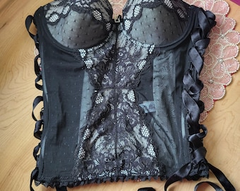 May include: Black corset with lace detailing and ribbon ties. The corset features a sweetheart neckline, lace cups, and a decorative lace panel down the front. Ruffles adorn the sides. The corset is displayed on a wooden surface.