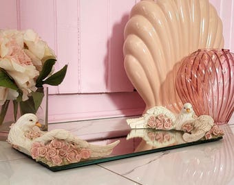 Może przedstawiać: Ozdobna lustrzana taca z dwiema figurkami białych gołębi, każda ozdobiona różowymi różami. Duży, ceramiczny element w kształcie muszli i różowy szklany wazon znajdują się w tle. Sceneria to pokój z różowymi ścianami i białą kafelkową podłogą.