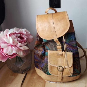 Puede incluir: Una mochila de cuero color canela con cierre de solapa y un pequeño bolsillo frontal. El cuerpo de la mochila presenta una tela estampada colorida con tonos de azul, verde y marrón. Tiene un asa en la parte superior.