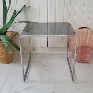 May include: A square side table with a smoked glass top and a chrome frame. The table is approximately 18 inches (46 cm) square and stands about 18 inches (46 cm) tall. The design is minimalist and modern.