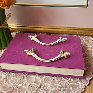 May include: Two decorative, curved drawer pulls with a white and gold finish, resting on a closed, magenta-colored book. The pulls have ornate, leaf-like details. A framed print and a bouquet of flowers are in the background.