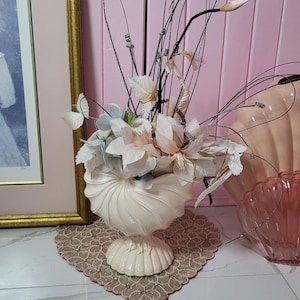 Peut inclure: Un vase à piédestal crème avec un motif de coquillage, rempli de fleurs artificielles blanches, pêche et bleues. L'arrangement comprend de longues tiges fines et est posé sur un napperon décoratif.