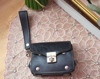 Könnte beinhalten: Eine kleine, schwarze Ledertasche mit silbernem Verschluss und Griff. Die Tasche ist rechteckig mit abgerundeten Ecken und silbernen Nieten. Sie liegt auf einem rosa Perlen-Tischset mit floralem Design. Im Hintergrund ein Strauß gelb-oranger Blumen.