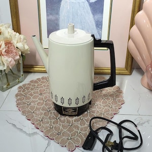 May include: A white ceramic mug with a black handle and the text "But First, Coffee" in a bold, black font. The mug is filled with a dark liquid, probably coffee, and rests on a light-coloured surface.