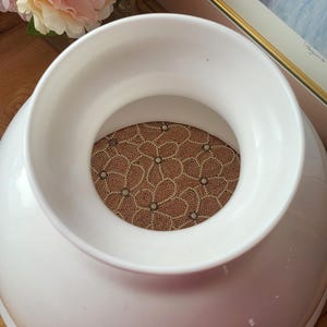 May include: A large, white ceramic vase with a wide opening and a decorative, patterned insert. The insert features a floral design in shades of brown and cream. The vase is set against a backdrop of flowers and framed artwork.