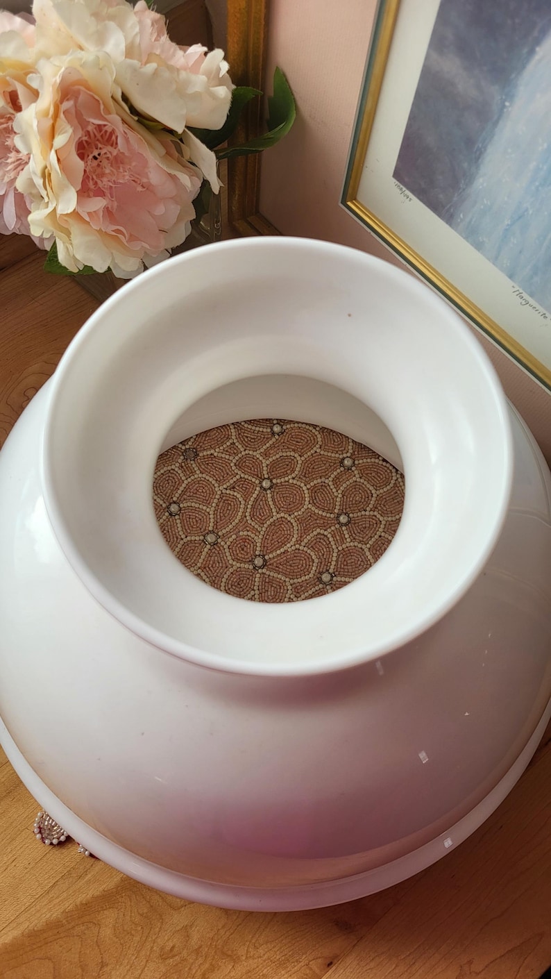 May include: A large, white ceramic vase with a wide opening and a decorative, patterned insert. The insert features a floral design in shades of brown and cream. The vase is set against a backdrop of flowers and framed artwork.