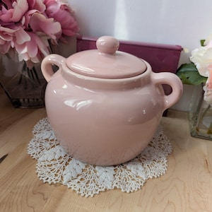 以下が含まれることがあります： ふた付きのピンクの陶器の壺で、持ち手が2つ付いています。壺は白いレースのコースターの上に置かれています。