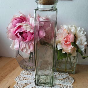 Puede incluir: Un tarro de vidrio transparente con tapa de corcho y diseño floral. El tarro está sobre un encaje blanco de ganchillo.