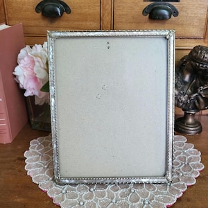 May include: A rectangular silver picture frame with a decorative border. The frame is empty, with a neutral-colored backing. It sits on a lace doily with a pink border. A pink flower arrangement and a bust are in the background.