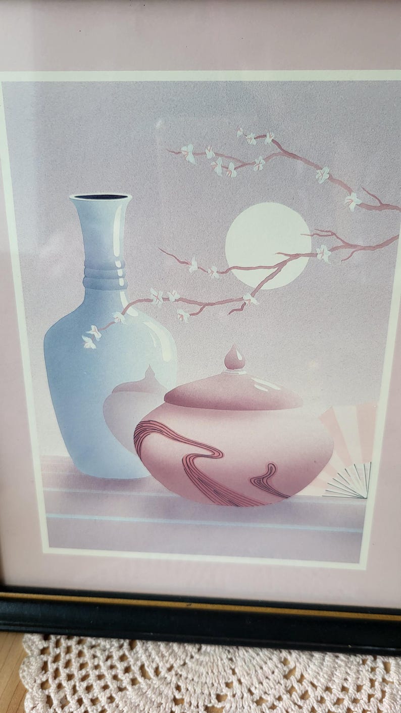 Op de afbeelding: Een ingelijste print van een stilleven met een blauwe vaas, een roze pot met deksel en een tak met witte bloesems tegen een lichtpaarse achtergrond.