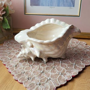 Può includere: Una fioriera a forma di conchiglia in ceramica bianca perlescente. La fioriera ha un bordo a balze e una superficie strutturata, appoggiata su un sottobicchiere rosa perlato. Una stampa incorniciata è sullo sfondo.