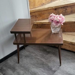Könnte beinhalten: Ein zweistufiger Holz-Beistelltisch mit brauner Oberfläche und schrägen Beinen. Der Tisch steht auf einem grauen Boden mit einem Strauß rosa Blumen in einer durchsichtigen Glasvase auf der oberen Ebene.