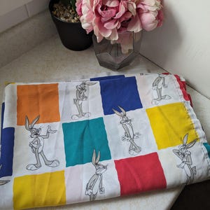 Könnte beinhalten: Ein weißer Stoff mit einem karierten Muster in Rot, Orange, Gelb, Blau, Grün und Türkis. Der Stoff zeigt ein sich wiederholendes Bild von Bugs Bunny in verschiedenen Posen.