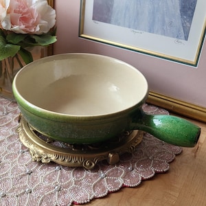 Puede incluir: Un cuenco de cerámica verde con asa, sobre un soporte decorativo de metal dorado. El cuenco tiene un interior beige claro y un exterior verde oscuro. El asa es de color verde liso.