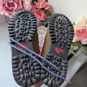 May include: A pair of brown leather hiking boots with black rubber soles and a red Hi-Tec logo. The boots are tied together with a purple shoelace.