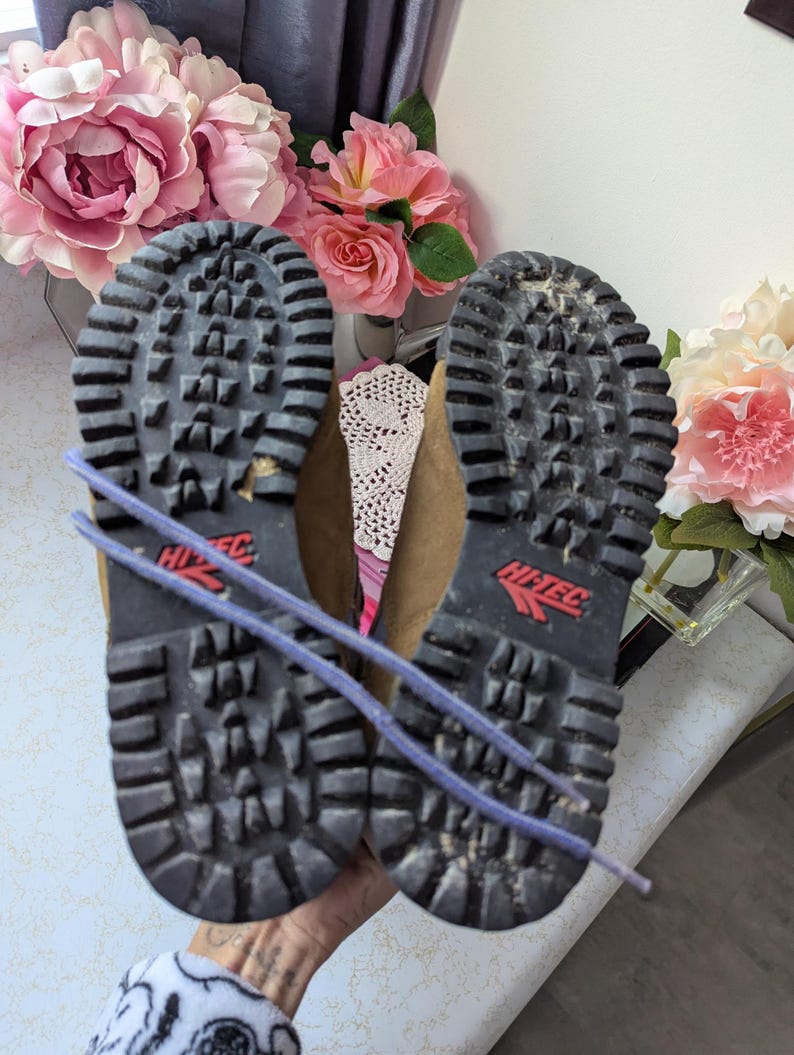 May include: A pair of brown leather hiking boots with black rubber soles and a red Hi-Tec logo. The boots are tied together with a purple shoelace.