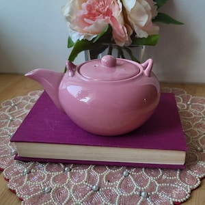 Peut inclure: Une théière en céramique rose avec un couvercle, posée sur un livre violet. Un bouquet de fleurs roses et blanches dans un vase en verre est derrière la théière. La théière a une forme arrondie et une finition brillante.