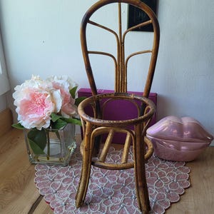 May include: A small, vintage bamboo chair with a round seat and back. The chair is brown and has a decorative design. A pink floral arrangement in a glass vase and a pink lip-shaped decorative object are nearby.
