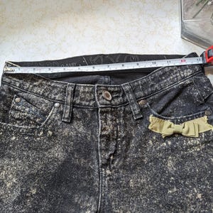 May include: Black acid wash denim jeans with a light brown bow detail on the right pocket. The waistband has the text "Baby Phat" printed on it.