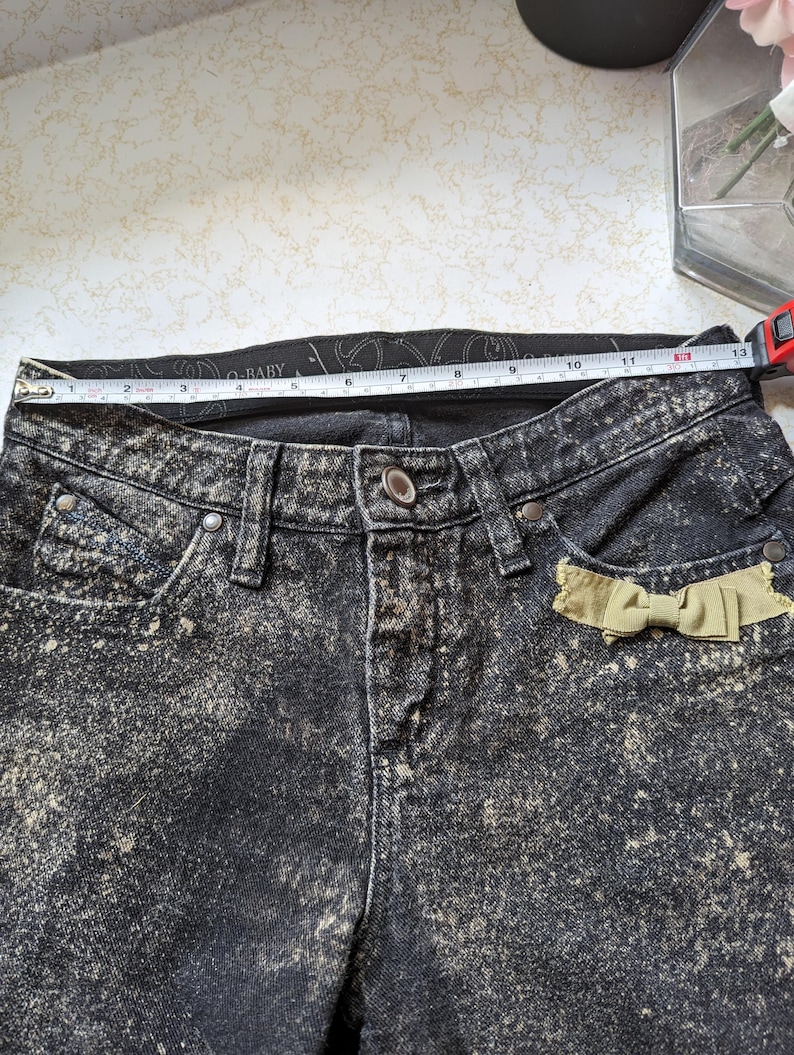 May include: Black acid wash denim jeans with a light brown bow detail on the right pocket. The waistband has the text "Baby Phat" printed on it.