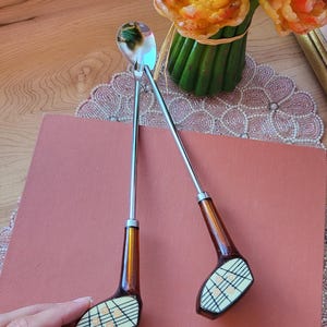 Könnte beinhalten: Zwei dekorative Löffel in Form von Golfschlägern mit silbernen Stielen und braunen Griffen. Die Löffelköpfe sind weiß mit einem Gittermuster und orangefarbenen Akzenten. Die Löffel liegen auf einem rosa Buch, im Hintergrund ein Strauß gelber Blumen.