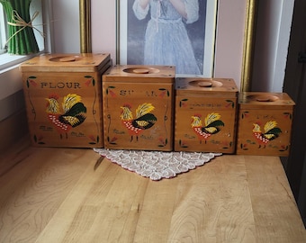 May include: A set of four wooden kitchen canisters with lids, featuring hand-painted rooster designs in red, yellow, and green. The canisters are labeled "FLOUR", "SUGAR", and "COFFEE". A framed portrait and decorative flowers are in the background.