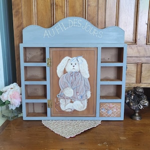 May include: A wooden wall shelf painted light blue with the text "AU FIL DES JOURS". The centre door features a painting of a bunny wearing a dress. The shelf has multiple compartments.