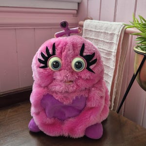 May include: A plush toy featuring bright pink faux fur, large blue eyes with long black eyelashes, and a purple heart-shaped patch on the front. The creature has a small purple antenna on top of its head and purple feet at the base.
