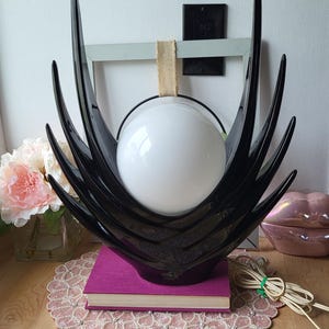 May include: A black, sculptural table lamp with a white globe light. The lamp has a stylized, spiky design and sits on a purple book. The lamp is on a wooden surface with a pink beaded placemat.
