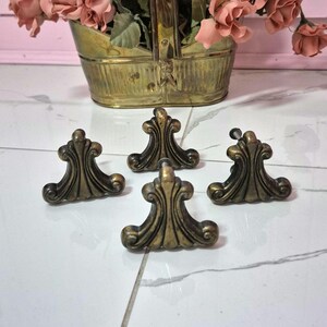 May include: Four antique brass drawer pulls featuring a decorative fan-like shape with curved edges and a dark, weathered finish. Each piece has a central mounting screw visible on the back, showcasing a symmetrical design with vertical ridges.