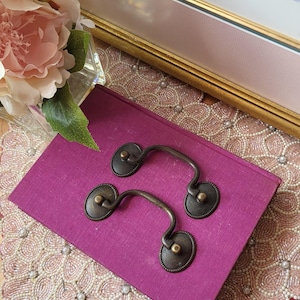 Peut inclure: Deux poignées de tiroir antiques en bronze avec une poignée incurvée, posées sur un livre violet. Les poignées ont une plaque arrière circulaire avec des détails décoratifs. Une œuvre d'art encadrée et un arrangement floral sont en arrière-plan.