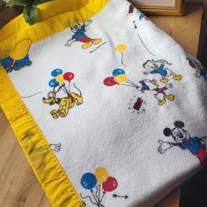 Puede incluir: Manta de bebé blanca con Mickey Mouse, Pluto y el Pato Donald con globos. La manta tiene un borde de satén amarillo. La manta está doblada sobre una superficie de madera.