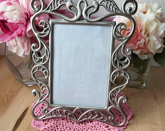 May include: A silver metal picture frame with an ornate design featuring swirls and leaves. The frame is sitting on a pink crocheted doily.