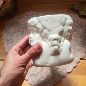 May include: A white ceramic planter featuring a sculpted design of cherubic faces. The planter has a rectangular shape with a wide opening. The design is detailed and the ceramic has a smooth finish. The planter is held by a hand.