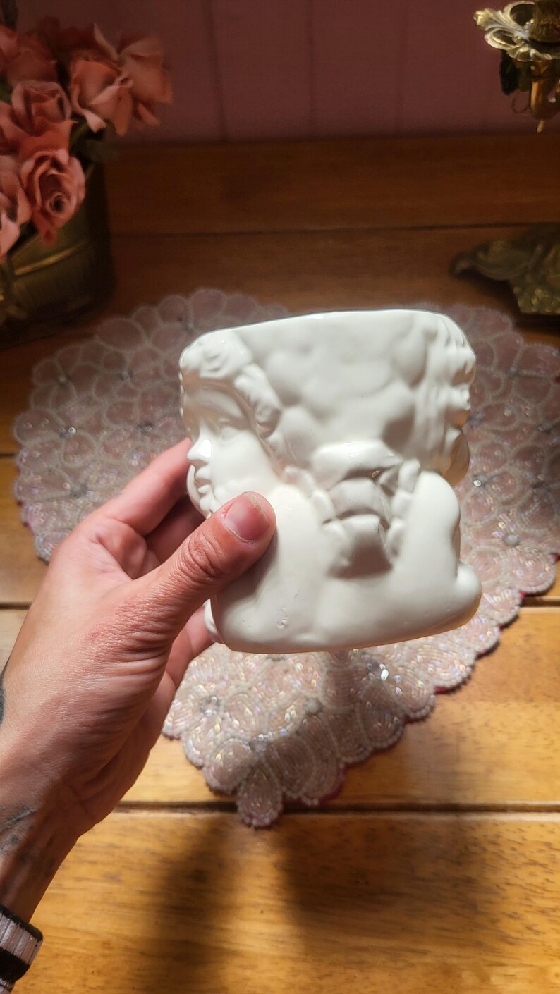 May include: A white ceramic planter featuring a sculpted design of cherubic faces. The planter has a rectangular shape with a wide opening. The design is detailed and the ceramic has a smooth finish. The planter is held by a hand.