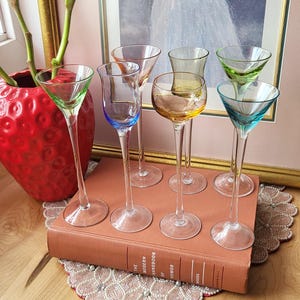 May include: Six stemmed glasses with coloured bowls in shades of green, blue, orange, and yellow. The glasses are displayed on a book, resting on a decorative doily. The glasses are ideal for serving drinks.