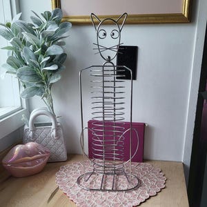 May include: A silver metal CD rack shaped like a cat with a pink heart-shaped beaded placemat on a wooden surface.