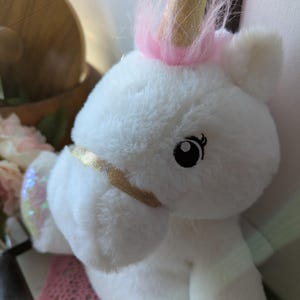 May include: White plush unicorn toy with a pink mane and a gold horn. The unicorn has a sparkly iridescent wing and black eyes.