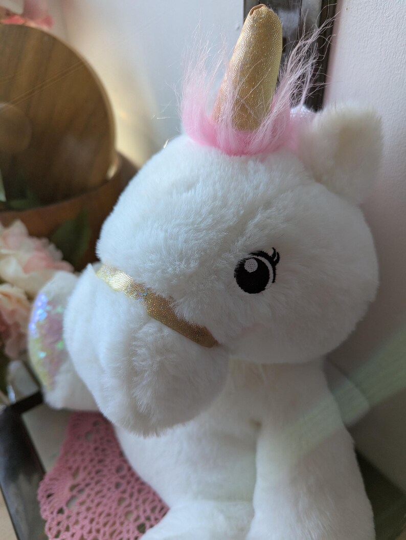 May include: White plush unicorn toy with a pink mane and a gold horn. The unicorn has a sparkly iridescent wing and black eyes.